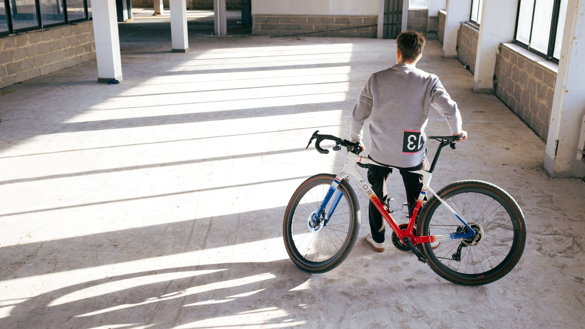 Cyclist standing next to a bicycle on a concrete floor with large windows in the background.