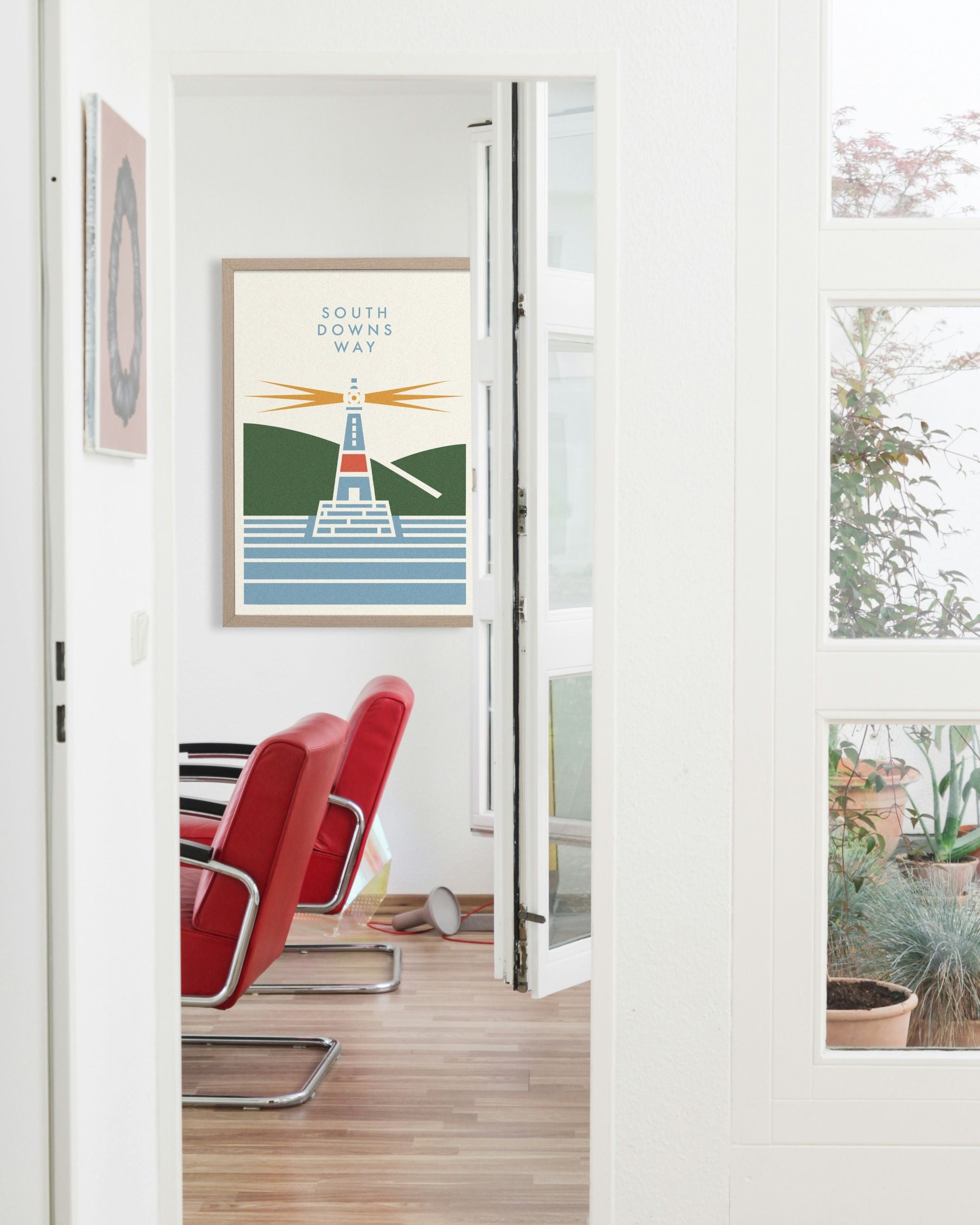 Framed South Down Way cycling artwork designed by Handmade Cyclist on a wall with red chairs and a glass door in the background.