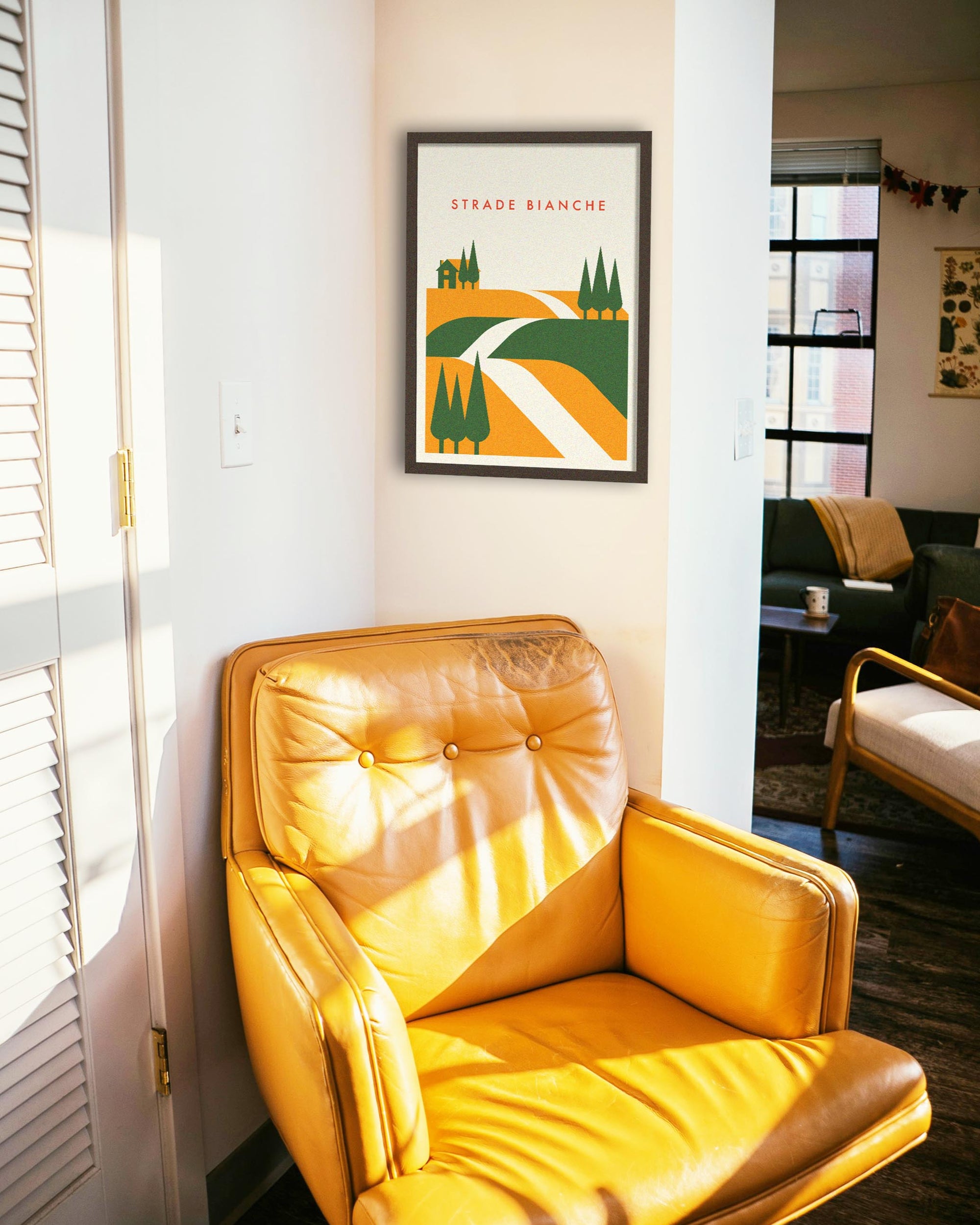 Yellow armchair in a room with a framed poster of Strade Bianchi on the wall, designed by the Handmade Cyclist
